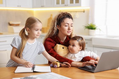 Photo of Single mother working with laptop and her daughters at table in kitchen