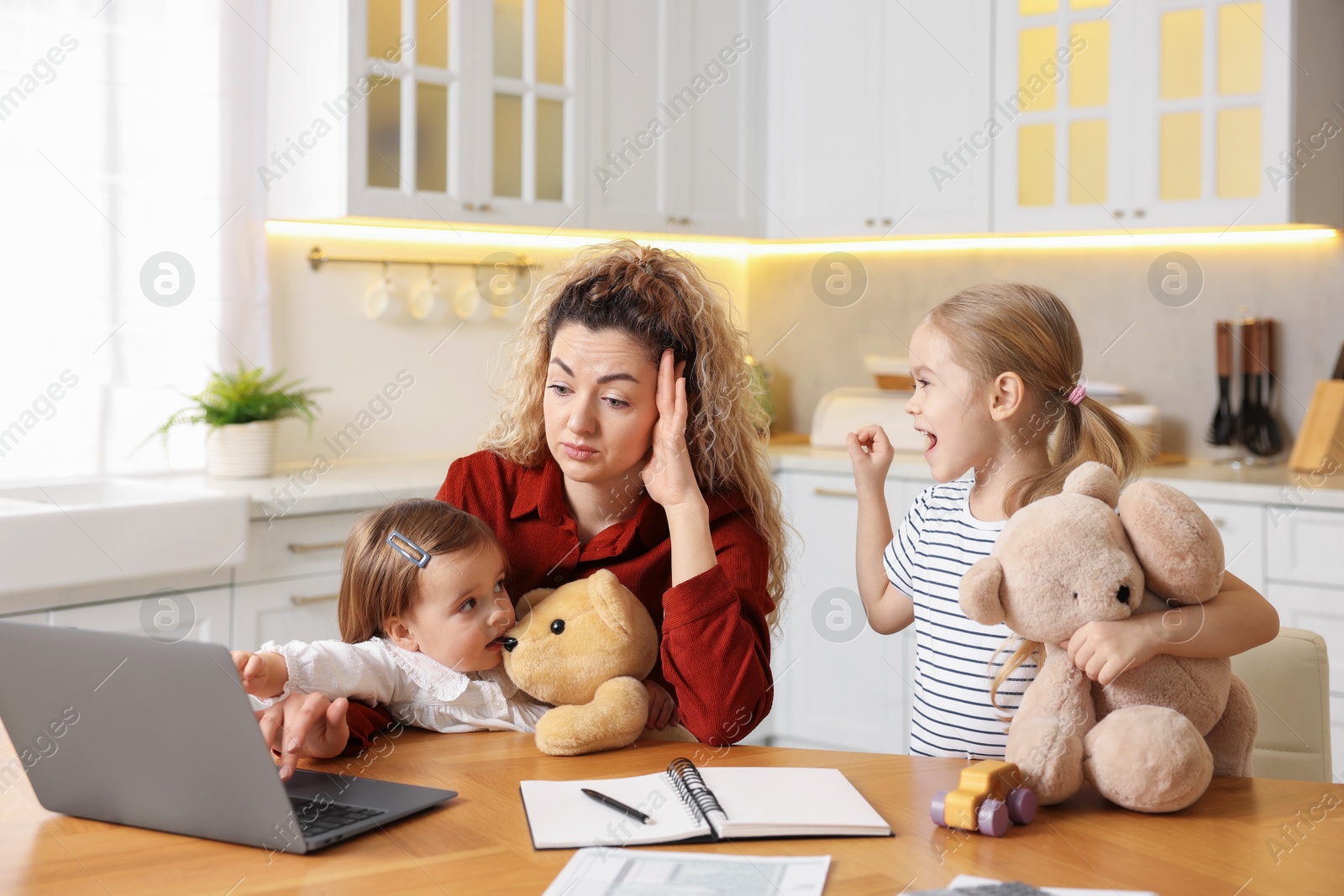 Photo of Single mother working with laptop while her daughters playing at table in kitchen