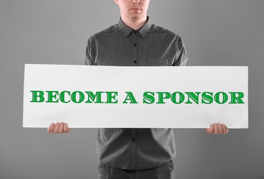 Man holding sign with text Become A Sponsor on grey background, closeup