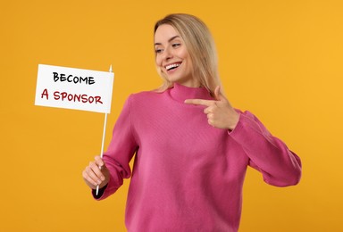 Happy young woman pointing at white flag with text Become A Sponsor on orange background