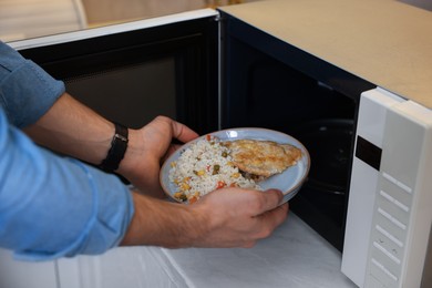 Photo of Man putting plate with lunch into microwave in kitchen, closeup