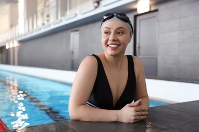 Photo of Happy woman wearing cap, goggles and swimsuit in swimming pool indoors