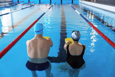 Young man and woman with kickboards swimming in pool indoors, back view