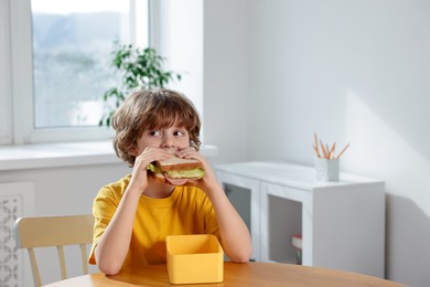 Cute little boy eating sandwich at wooden table in school. Space for text