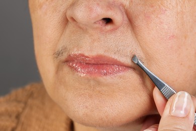 Photo of Senior woman plucking her mustache with tweezers on grey background, closeup