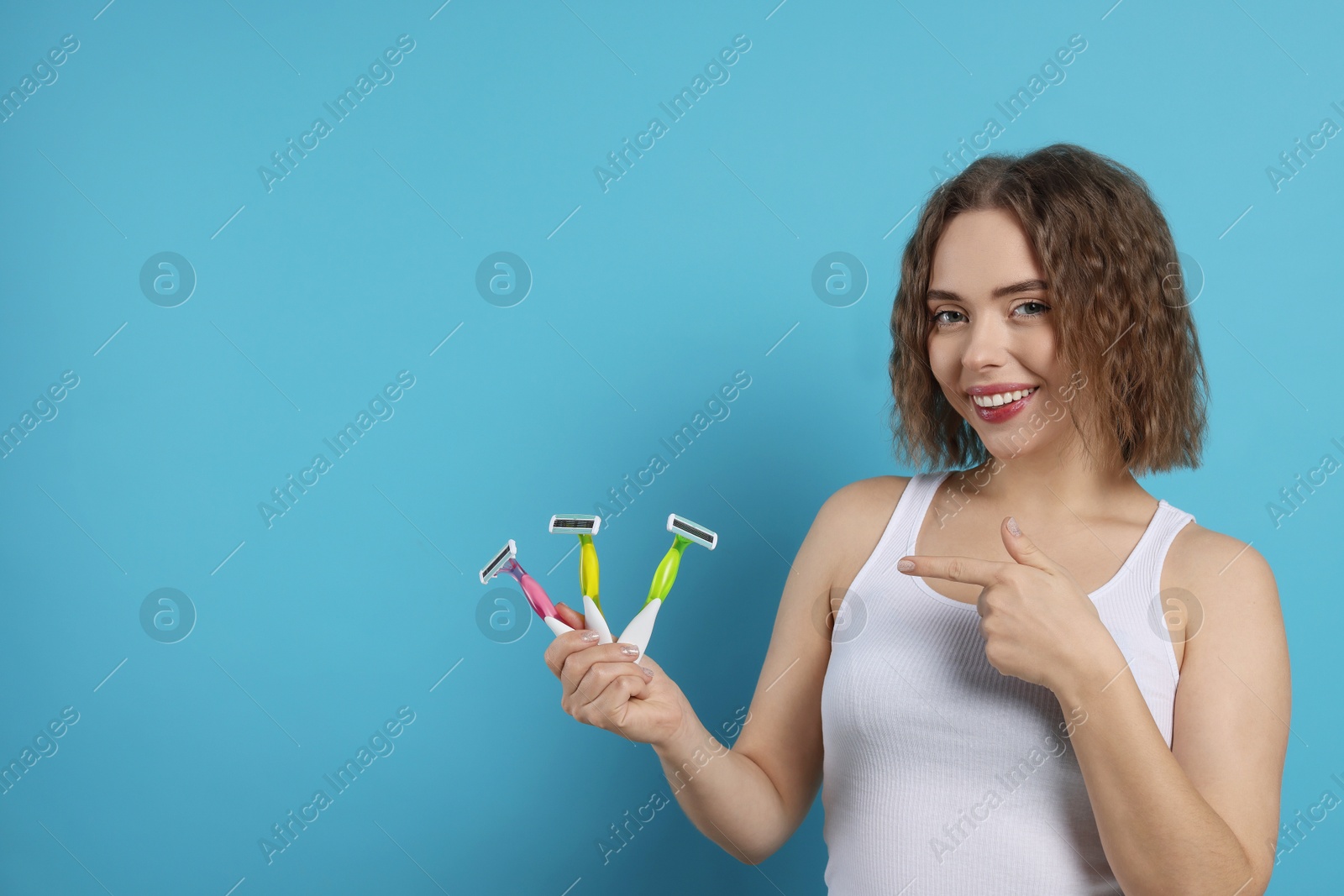 Photo of Smiling woman pointing at different razors on light blue background, space for text. Hair removal tools