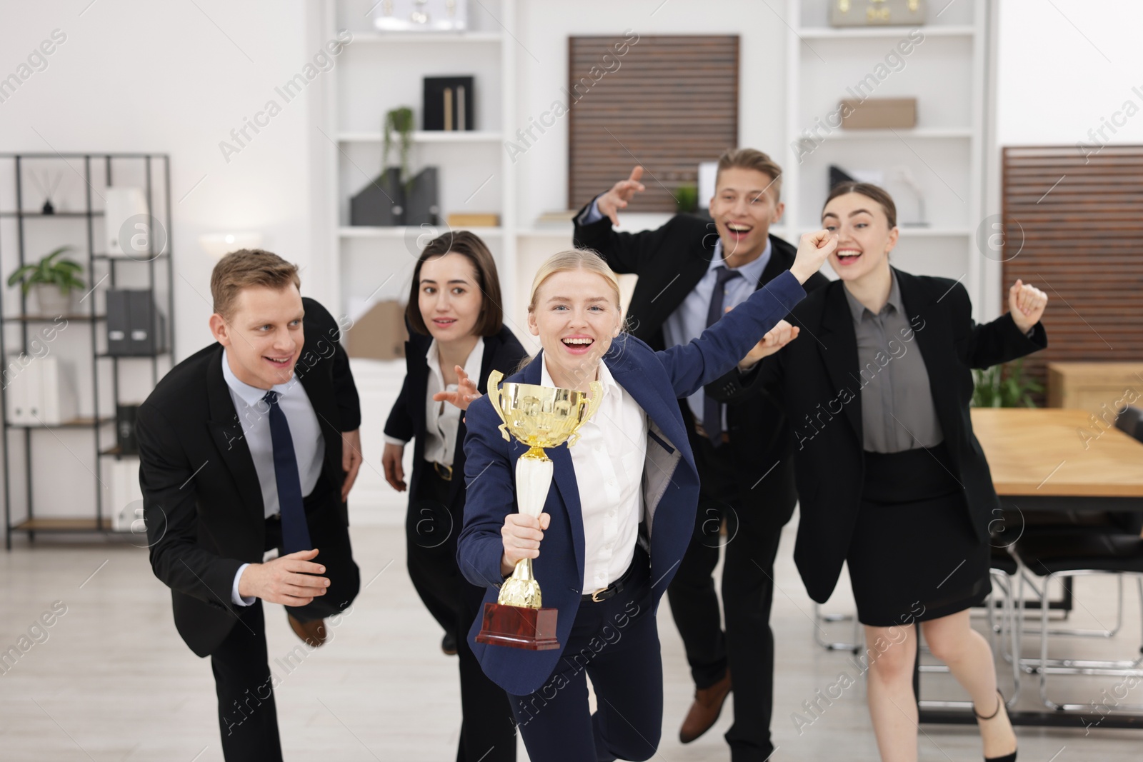 Photo of Competition concept. Happy businesswoman with trophy cup running ahead of her colleagues in office