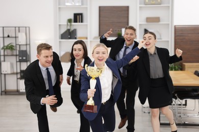 Competition concept. Happy businesswoman with trophy cup running ahead of her colleagues in office