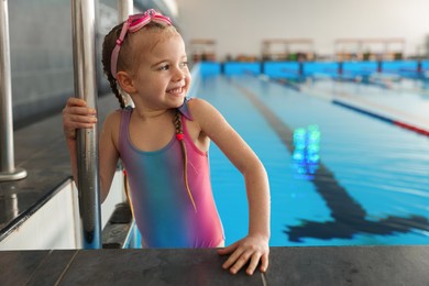 Little girl with goggles getting out of swimming pool indoors, space for text