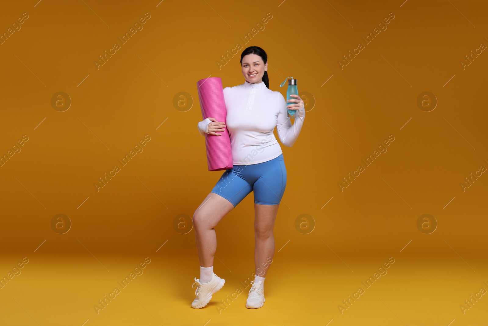 Photo of Plus size woman in gym clothes with water bottle and fitness mat on orange background