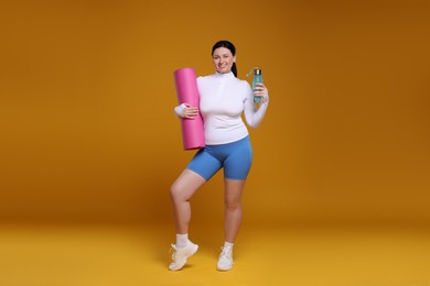 Plus size woman in gym clothes with water bottle and fitness mat on orange background