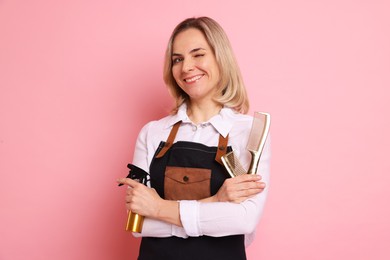 Smiling hairdresser with combs and spray bottle on pink background