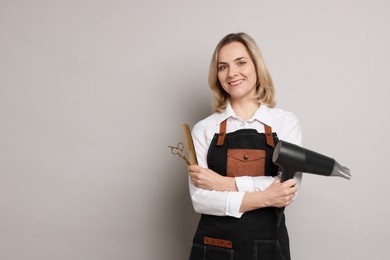 Smiling hairdresser with dryer, scissors and comb on gray background, space for text