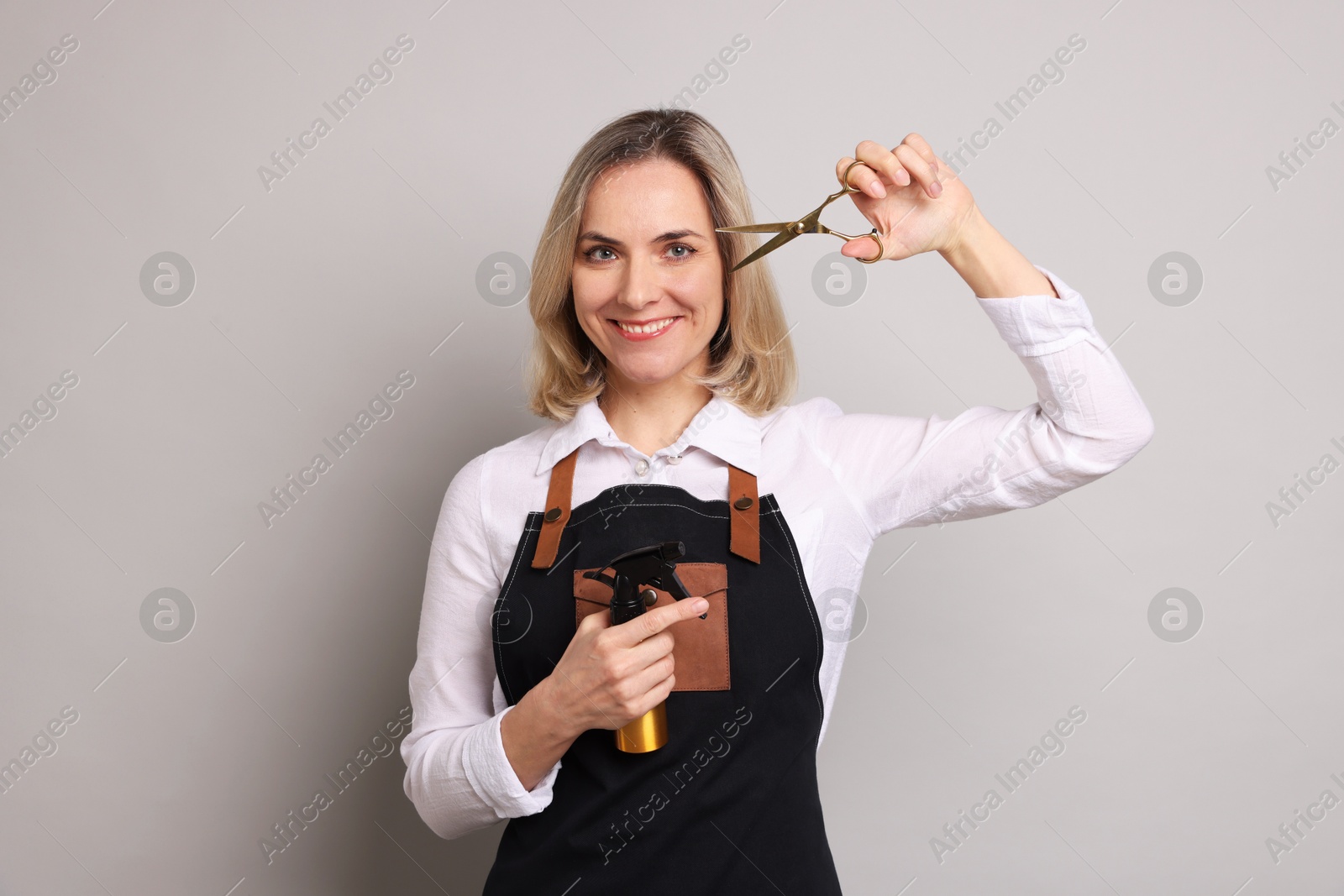 Photo of Smiling hairdresser with scissors and spray bottle on gray background