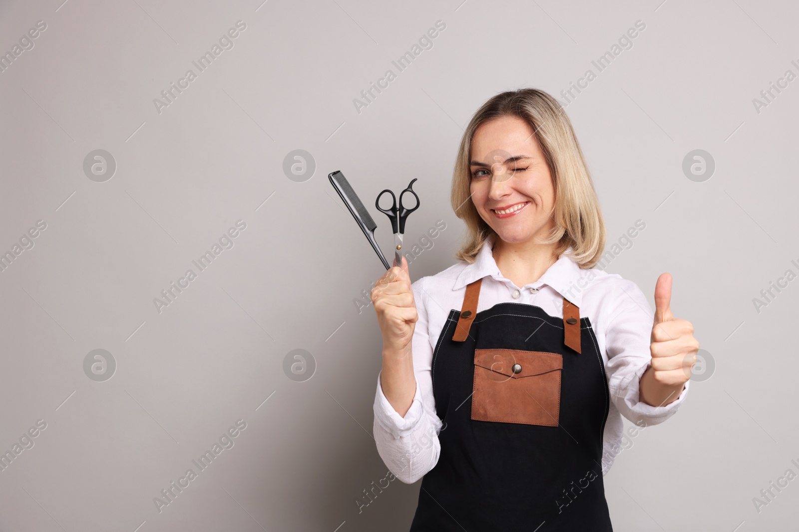 Photo of Smiling hairdresser with comb and scissors showing thumbs up on gray background, space for text