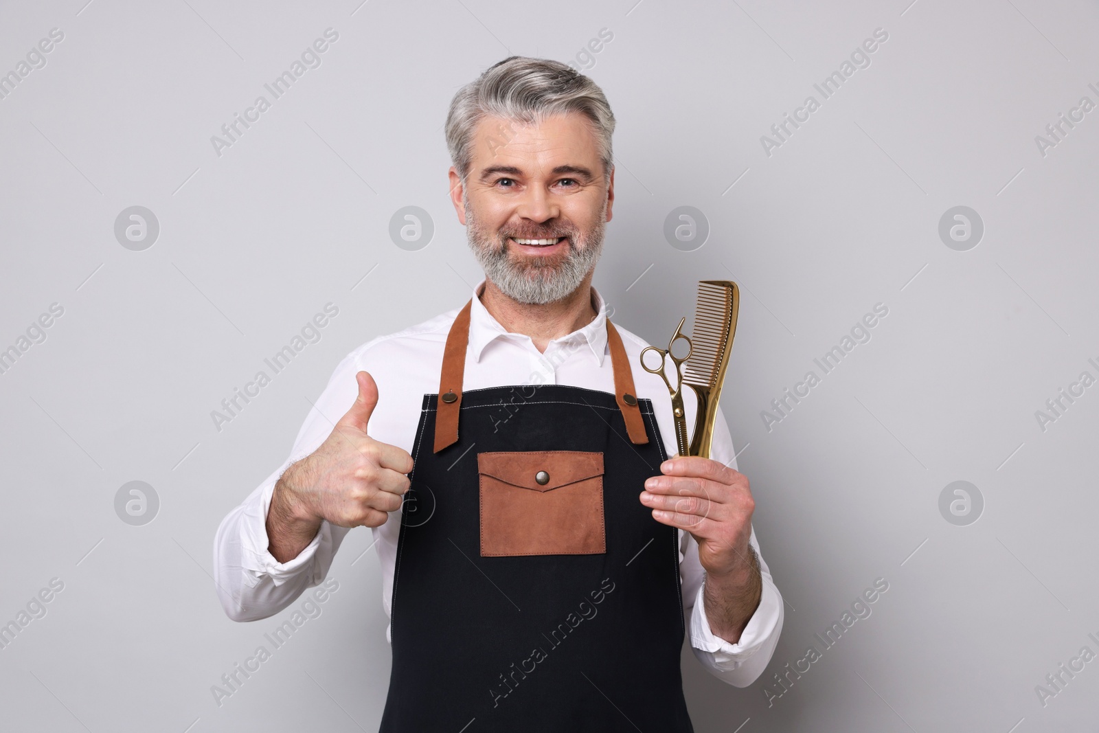 Photo of Smiling hairdresser with scissors and comb showing thumbs up on gray background