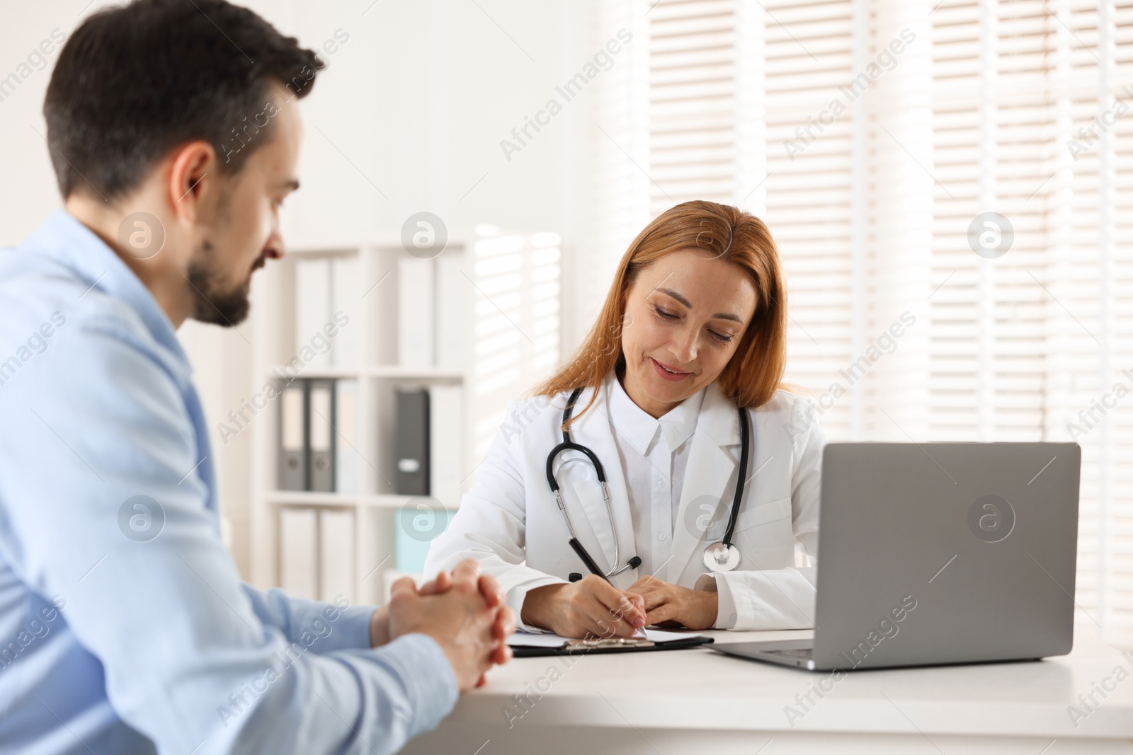 Photo of Cardiologist taking notes while consulting patient at table in clinic