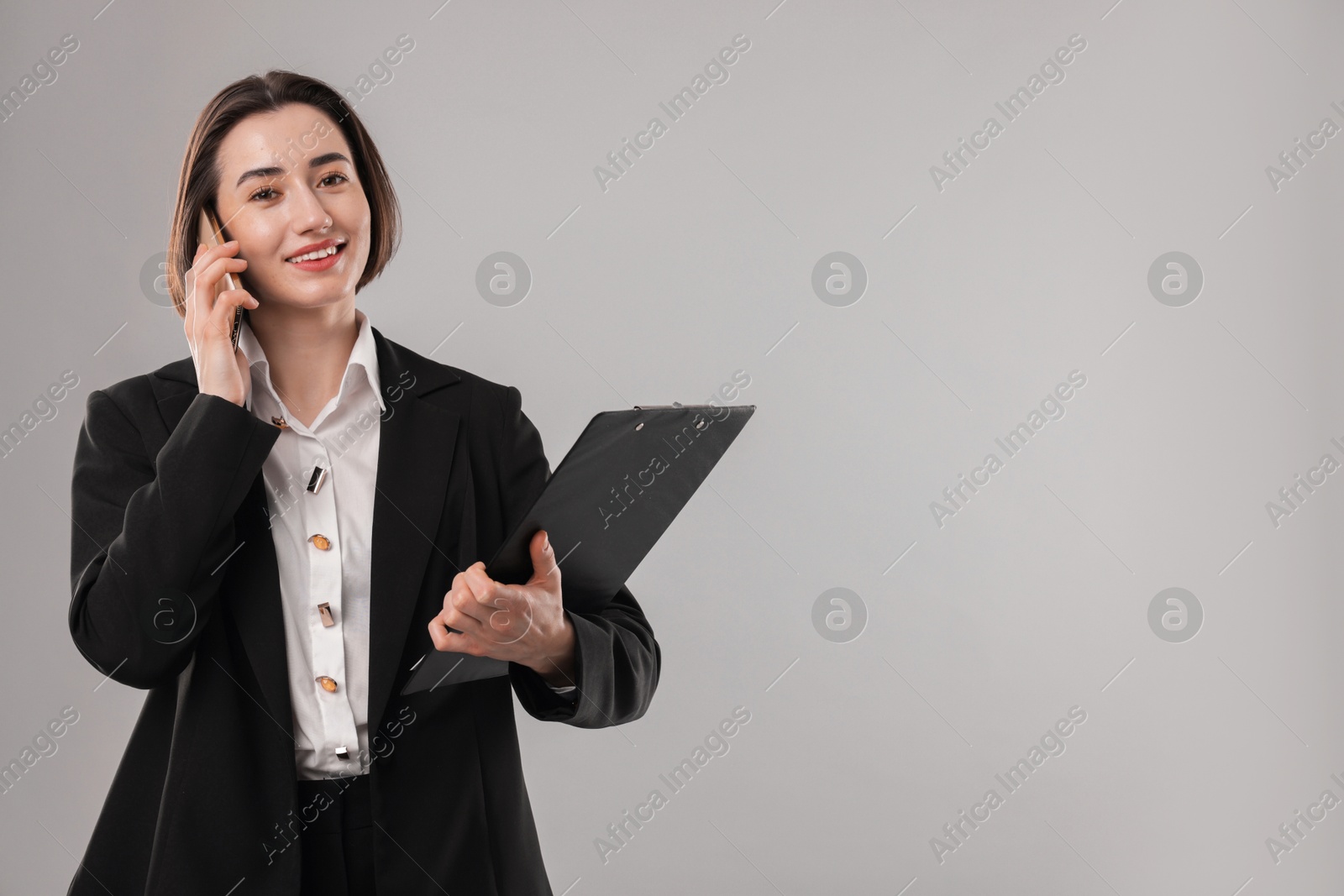 Photo of Smiling secretary with clipboard talking on smartphone against grey background