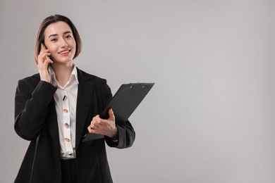 Smiling secretary with clipboard talking on smartphone against grey background