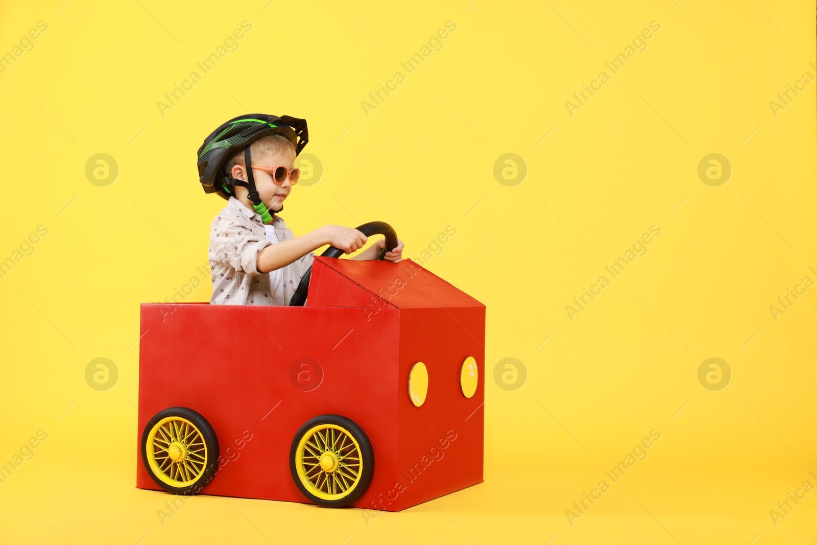 Photo of Little boy driving car made of cardboard on yellow background. Space for text