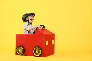 Little boy driving car made of cardboard on yellow background. Space for text