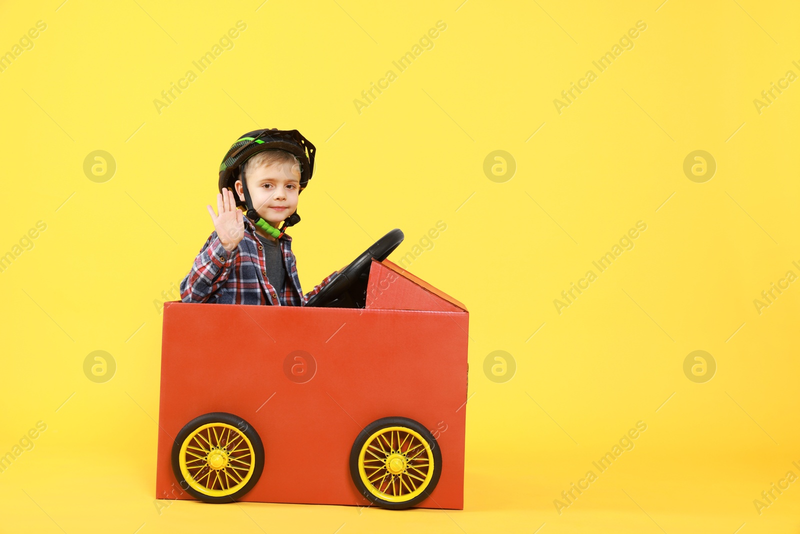 Photo of Little boy waving while driving car made of cardboard on yellow background. Space for text