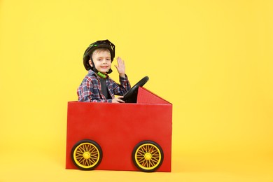 Photo of Little boy waving while driving car made of cardboard on yellow background. Space for text