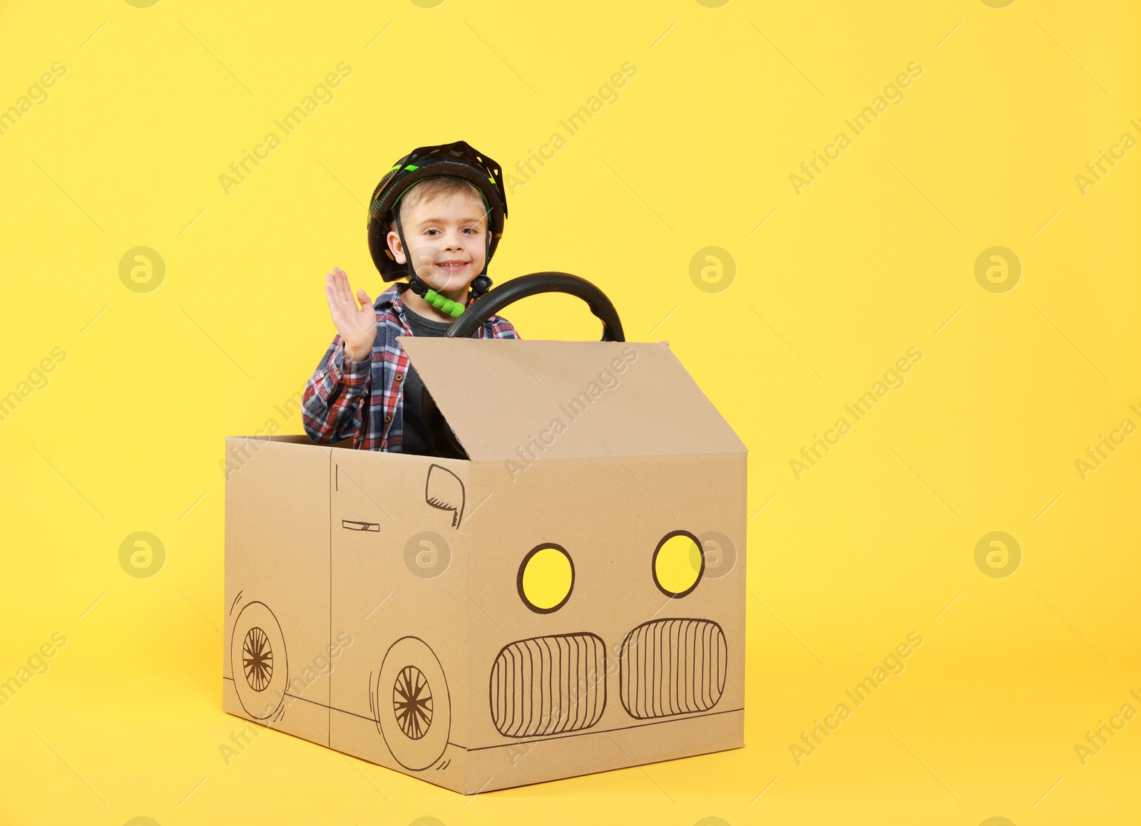 Photo of Little boy waving while driving car made of cardboard on yellow background. Space for text