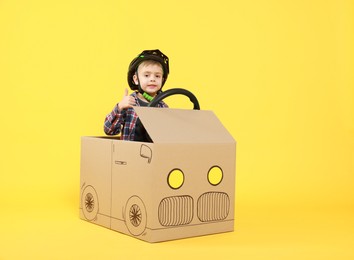 Little boy showing thumbs up while driving car made of cardboard on yellow background. Space for text