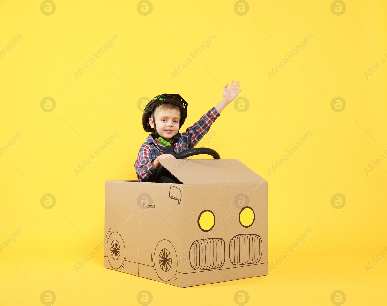 Photo of Little boy waving while driving car made of cardboard on yellow background