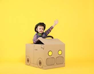 Little boy waving while driving car made of cardboard on yellow background