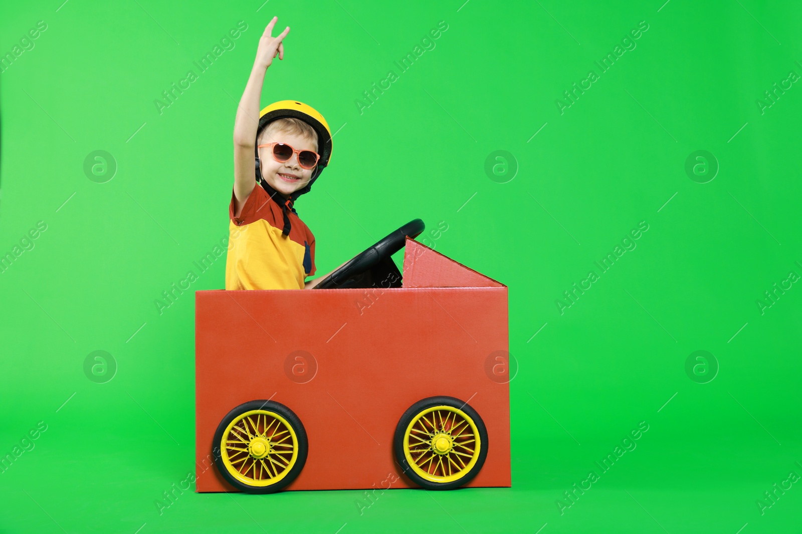 Photo of Little boy showing sign of horns while driving car made with cardboard on green background. Space for text