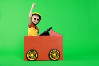 Little boy showing sign of horns while driving car made with cardboard on green background. Space for text