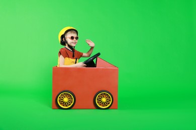 Little boy waving while driving car made of cardboard on green background. Space for text