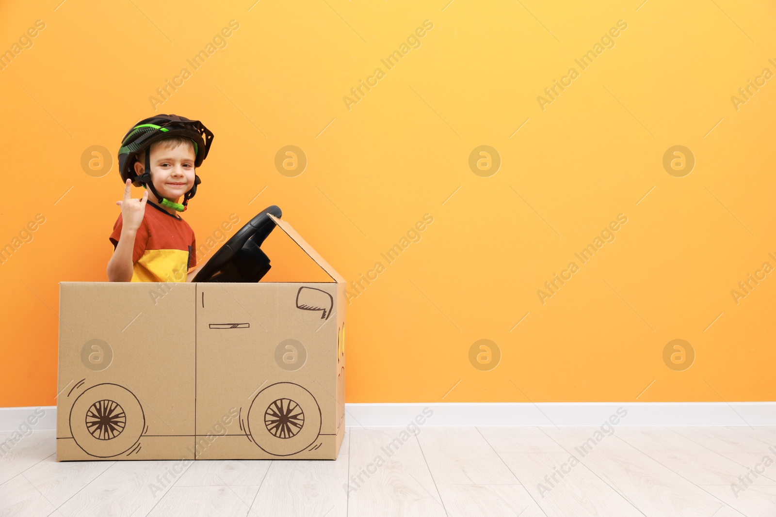 Photo of Little boy showing sign of horns while driving car made with cardboard against orange wall. Space for text