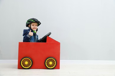 Photo of Little boy showing thumbs up while driving car made of cardboard against light wall. Space for text