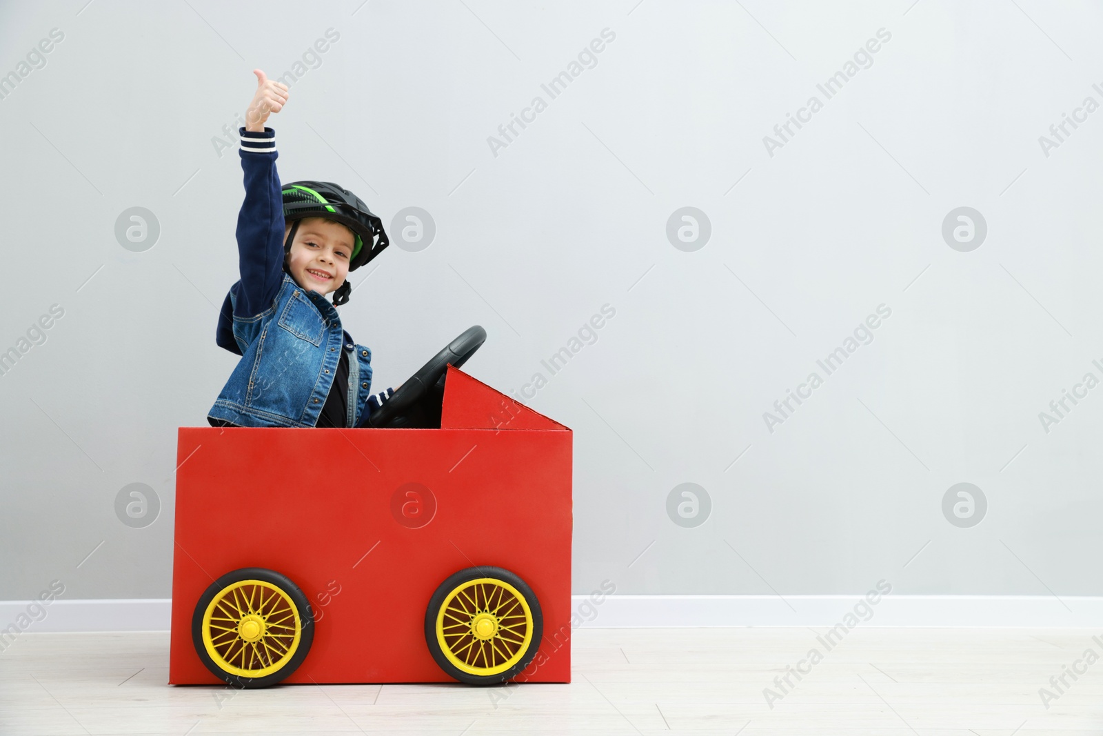 Photo of Little boy showing thumbs up while driving car made of cardboard against light wall. Space for text