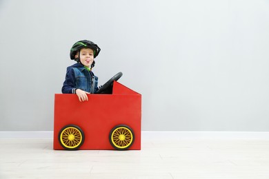 Photo of Little boy showing tongue while driving car made of cardboard against light wall. Space for text