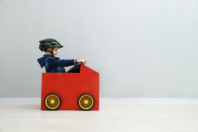 Photo of Little boy driving car made of cardboard against light wall. Space for text