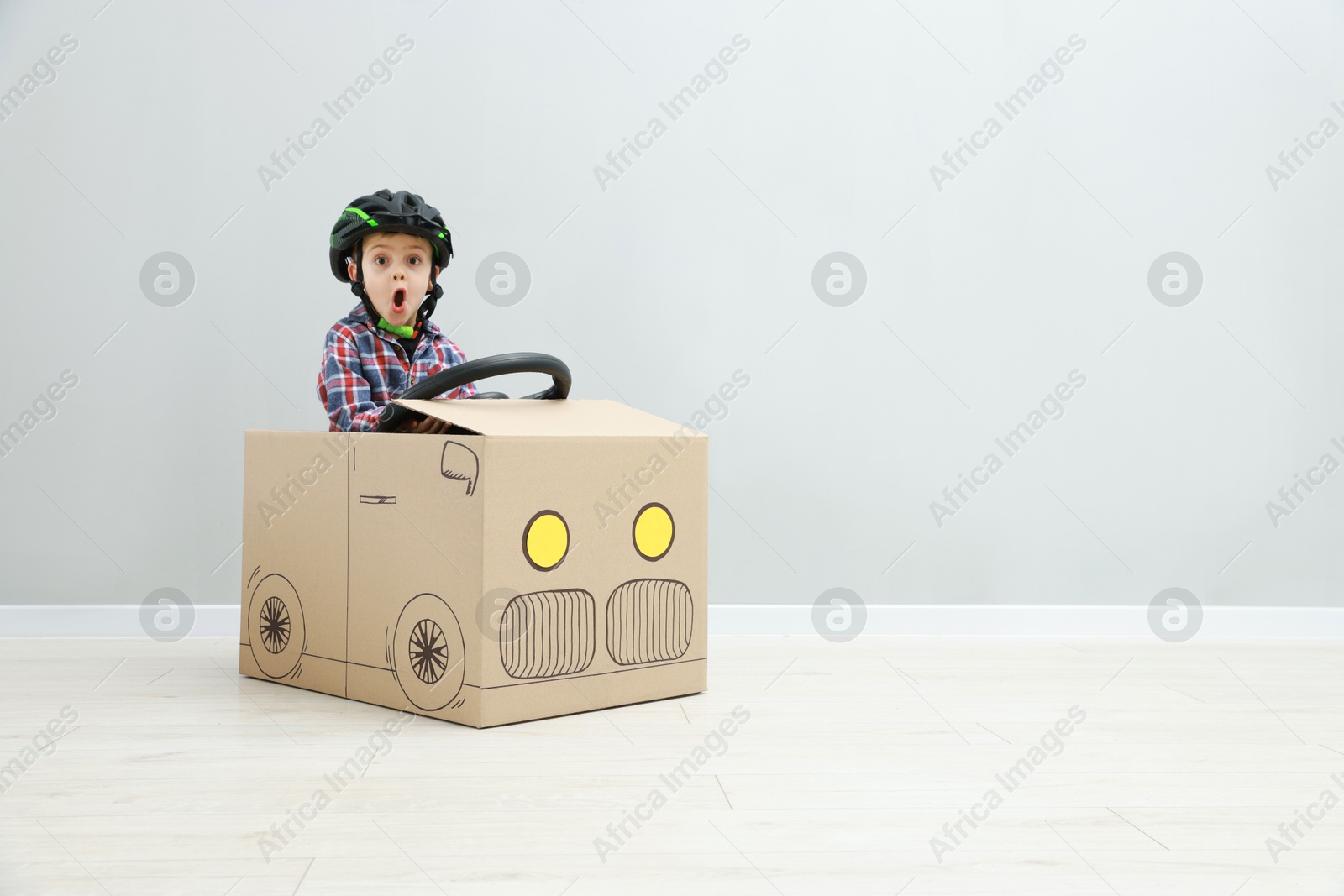 Photo of Surprised little boy driving car made of cardboard against light wall. Space for text