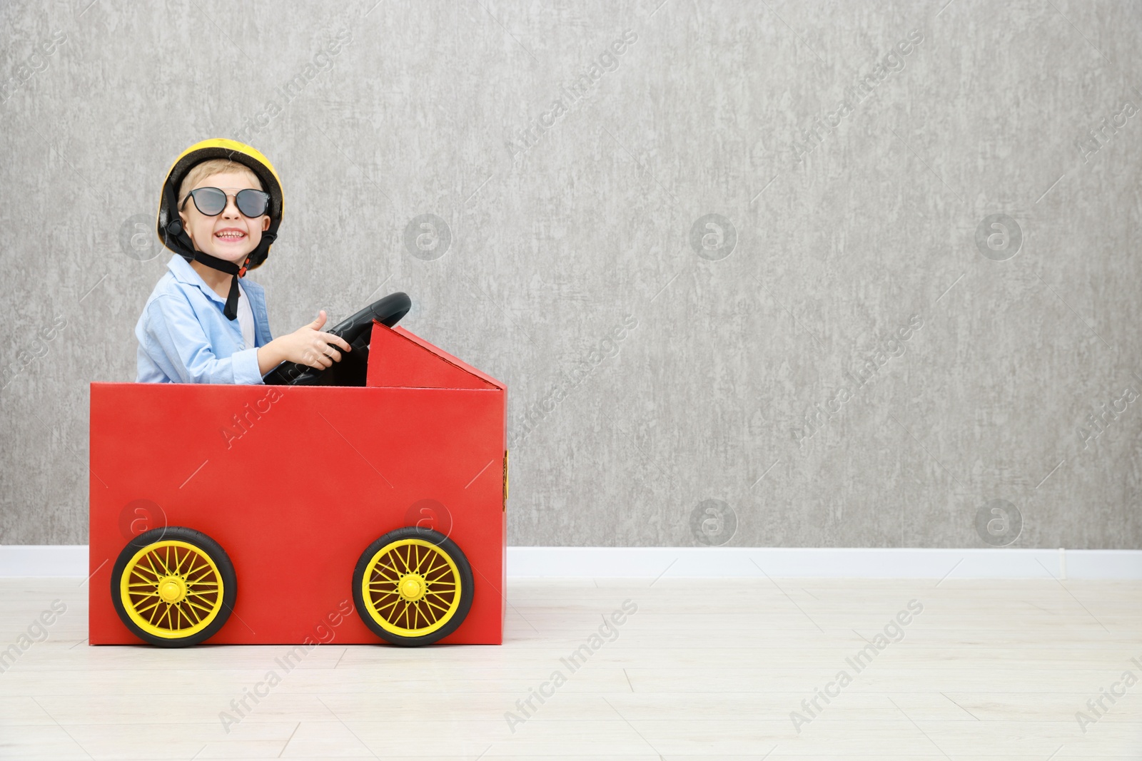 Photo of Little boy driving car made of cardboard against grey wall. Space for text