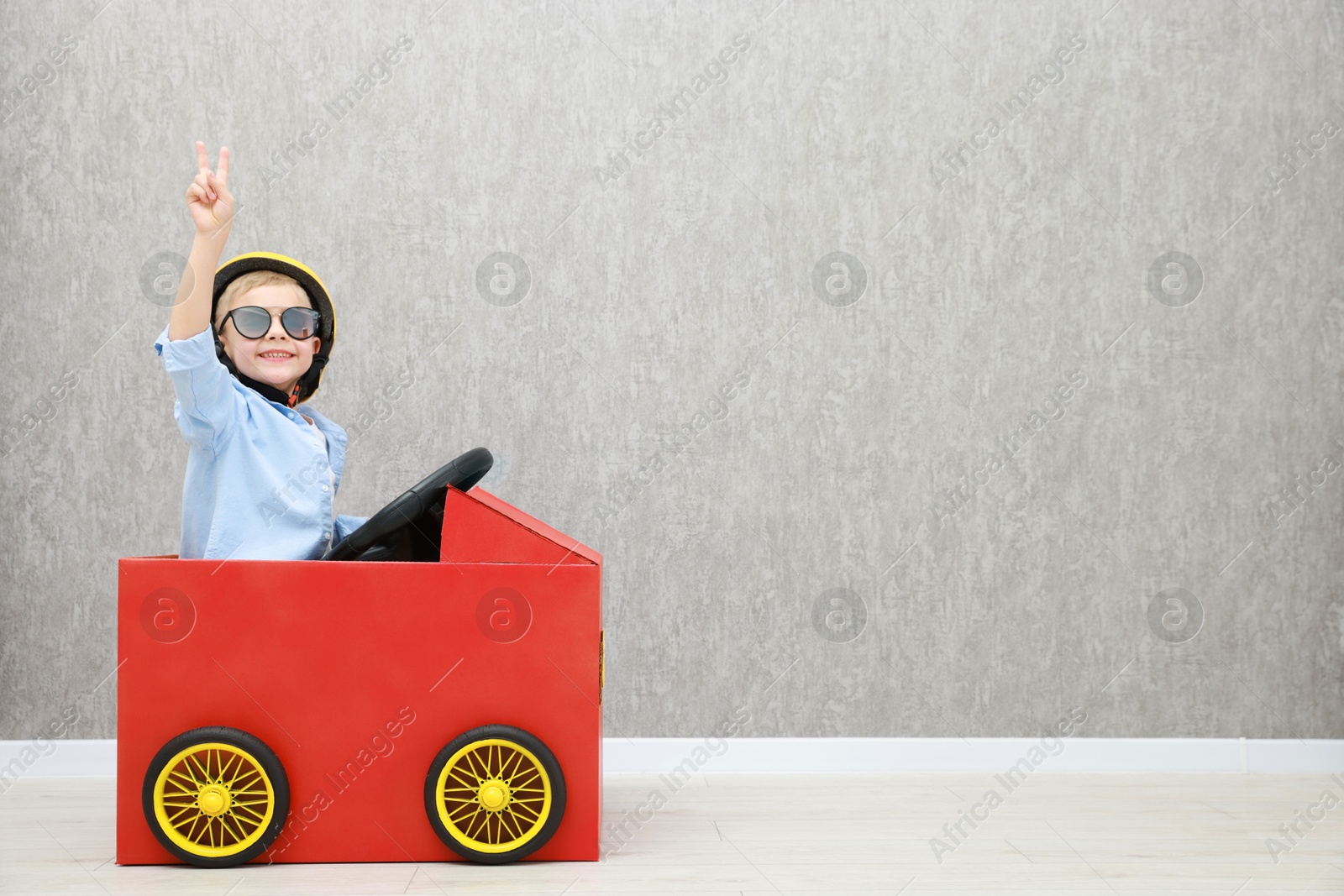 Photo of Little boy showing V-sign while driving car made of cardboard against grey wall. Space for text