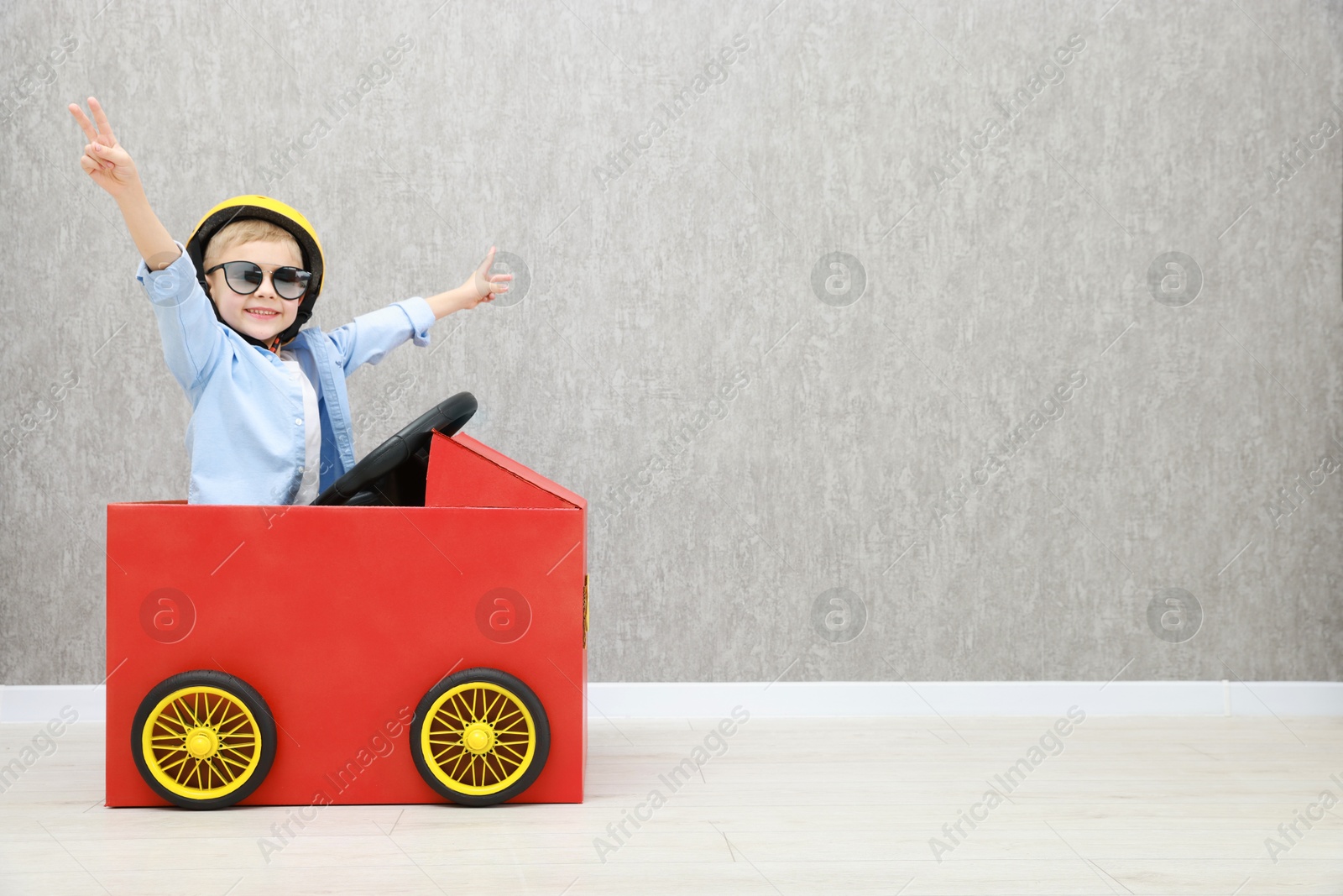 Photo of Little boy showing V-sign while driving car made of cardboard against grey wall. Space for text