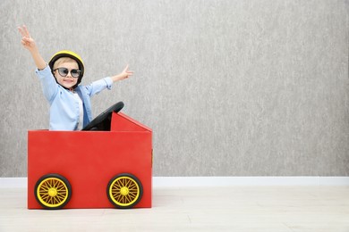 Little boy showing V-sign while driving car made of cardboard against grey wall. Space for text