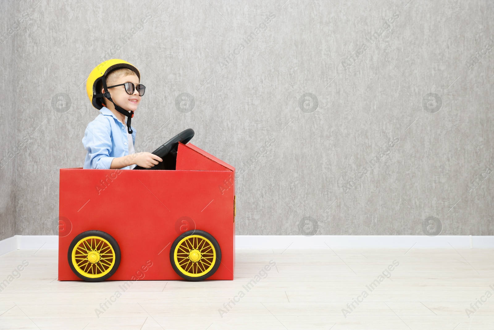 Photo of Little boy driving car made of cardboard against grey wall. Space for text