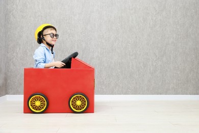 Little boy driving car made of cardboard against grey wall. Space for text