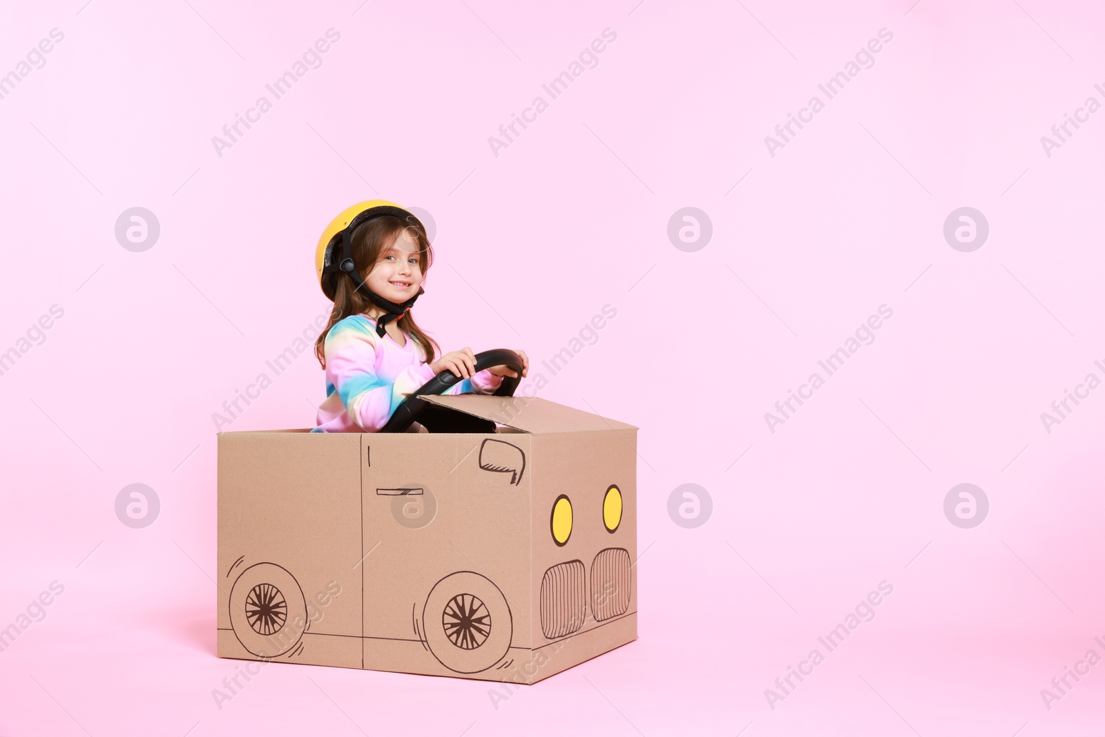 Photo of Little girl driving car made with cardboard on light pink background. Space for text