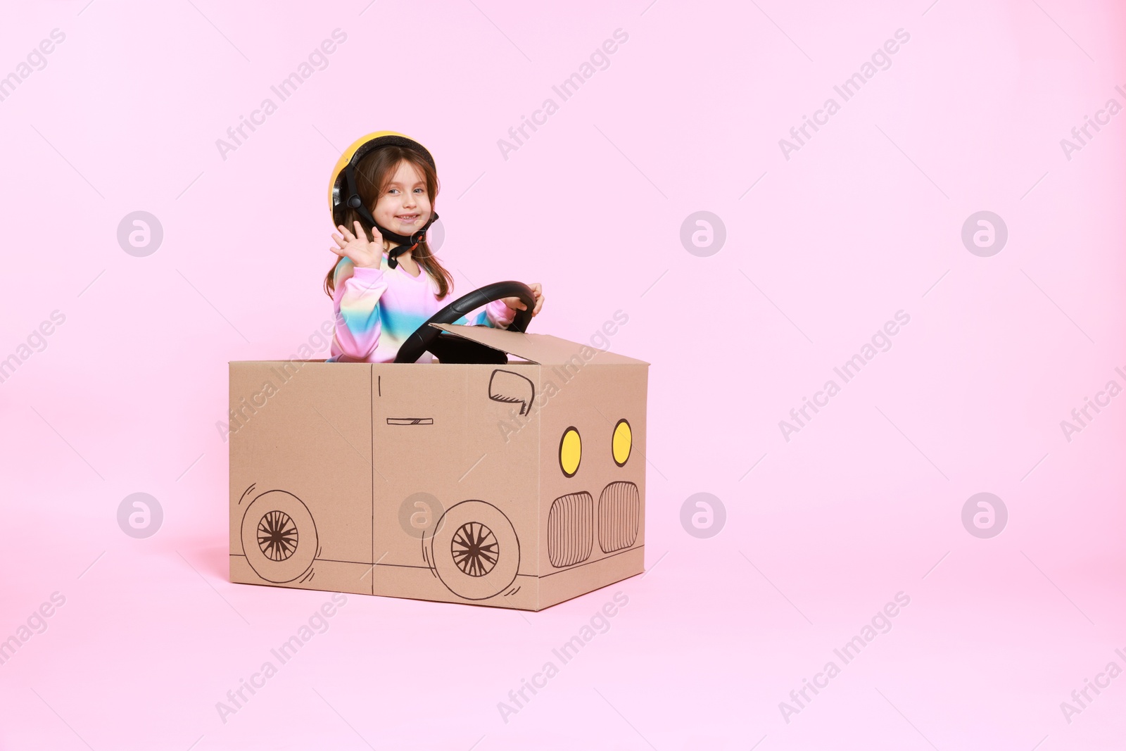 Photo of Little girl waving while driving car made with cardboard on light pink background