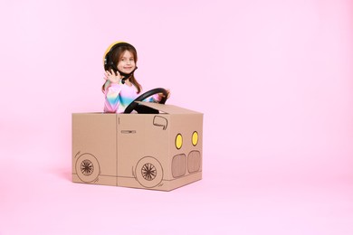 Photo of Little girl waving while driving car made with cardboard on light pink background