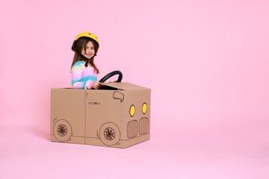 Photo of Little girl driving car made with cardboard on light pink background. Space for text